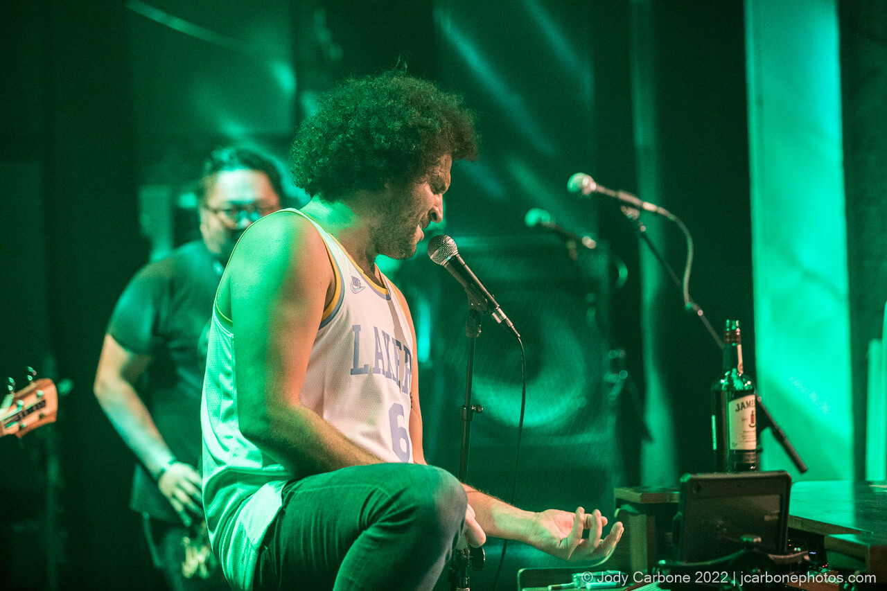 A concert photography portrait of Andy Frasco playing air guitar under green lights
